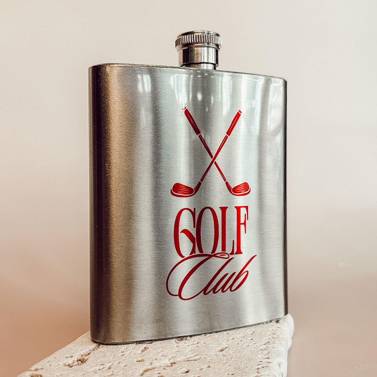 Edelstahl-Flachmann mit rotem Golf-Club-Motiv auf hellem Steinpodest